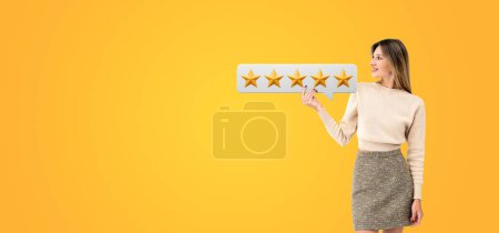 Photo for Smiling young woman holding up a speech bubble with five gold stars, empty orange background. Concept positive review, evaluation and customer satisfied - Royalty Free Image