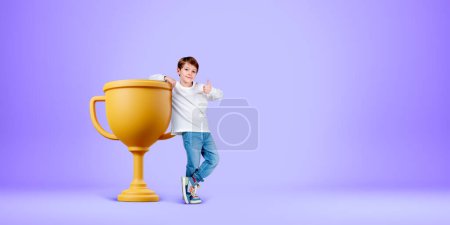 Téléchargez les photos : Child showing a thumb up, full length standing near big gold champion cup on light copy space purple background. Concept of first place, winner and success - en image libre de droit