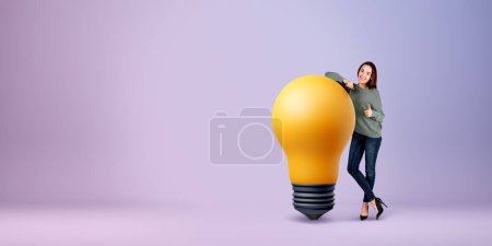 Téléchargez les photos : Femme souriante montrant le pouce levé, debout sur toute la longueur près d'une grande ampoule sur fond violet vide. Concept de soutien, créativité, plan et idée d'entreprise - en image libre de droit