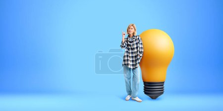 Téléchargez les photos : Femme doigt pointant vers le haut, pleine longueur debout près de grande ampoule sur fond bleu vide. Concept d'inspiration, start-up, créativité, connaissance et idée d'entreprise - en image libre de droit