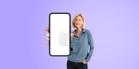 Foto de Mujer joven alegre en ropa casual que muestra el teléfono inteligente con pantalla simulada de pie sobre el fondo púrpura. Concepto de publicidad de aplicaciones y sitios web - Imagen libre de derechos