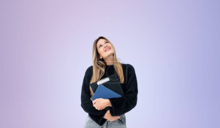 Photo for Portrait of thoughtful young woman university or college student holding notebooks and looking upwards standing over purple background. Concept of planning - Royalty Free Image