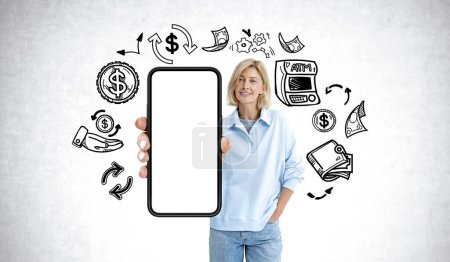 Photo for Smiling woman showing mockup blank smartphone display in hand, cashback doodle sketch with money and atm, grey concrete wall background. Concept of refund, online payment and service - Royalty Free Image