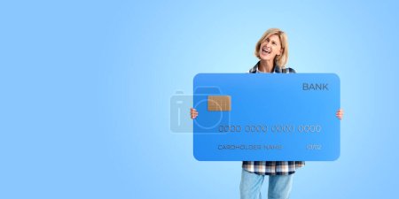 Téléchargez les photos : Joyeux sourire femme blonde tenant une carte bleue de crédit bancaire, copier l'espace fond vide. Concept de paiement, d'achat, de financement en ligne et de monnaie électronique - en image libre de droit