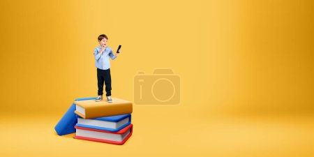 Téléchargez les photos : Petit garçon étonné avec smartphone debout sur une pile de livres sur fond jaune. Concept de bibliothèque numérique et d'enseignement en ligne, e-learning - en image libre de droit