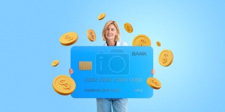 Foto de Joven mujer europea sosteniendo una gran tarjeta de crédito con monedas de dólar alrededor de su pie sobre fondo azul. Concepto de banca en línea y reembolso - Imagen libre de derechos