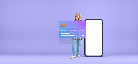 Foto de Gritando mujer feliz sosteniendo una gran maqueta de tarjeta de crédito, pantalla de teléfono en blanco sobre fondo púrpura vacío. Concepto de compras en línea, pago y banca móvil - Imagen libre de derechos
