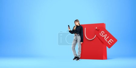 Téléchargez les photos : Femme concentrée regardant le téléphone debout près du grand sac à provisions rouge, copier l'espace fond bleu. Concept de shopping en ligne, bonne offre, site web et publicité - en image libre de droit