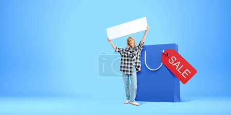 Foto de Mujer gritando feliz mostrando un letrero en blanco maqueta, de pie cerca de una gran bolsa de compras con etiqueta de venta en el fondo azul vacío. Concepto de compras en línea y ganga - Imagen libre de derechos