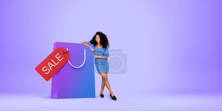 Photo for Beautiful young African American woman standing near big shopping bag with sale tag over purple copy space background. Concept of shopping - Royalty Free Image