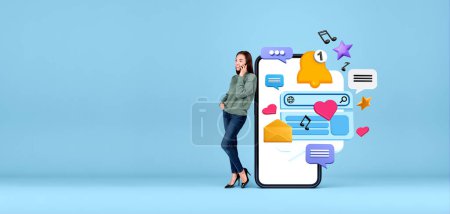 Téléchargez les photos : Jeune femme européenne parlant au téléphone debout près d'un grand smartphone avec des icônes de médias sociaux sur fond d'espace de copie bleue. Concept de communication en ligne - en image libre de droit