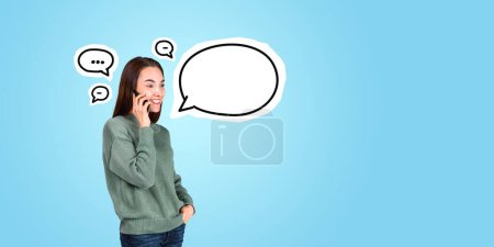 Foto de Retrato de una joven alegre hablando en un teléfono inteligente con burbujas de habla alrededor de ella de pie sobre el fondo del espacio de copia azul. Concepto de comunicación - Imagen libre de derechos
