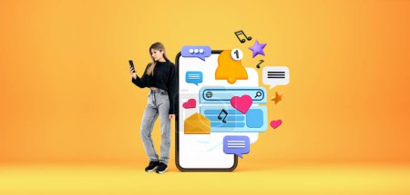 Photo for Young woman with smartphone and headphones standing near big smartphone with social media icons over yellow background. Concept of online communication - Royalty Free Image