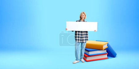 Téléchargez les photos : Femme souriante montrant un panneau de maquette, debout près de la pile de livres de dessins animés sur fond bleu. Concept d'éducation, publicité, cours et recommandation - en image libre de droit