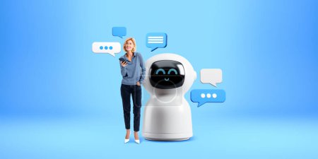 Téléchargez les photos : Jeune femme européenne avec smartphone debout près de grand robot blanc AI intelligence artificielle chat avec bulles de parole sur fond bleu. Concept d'apprentissage automatique - en image libre de droit