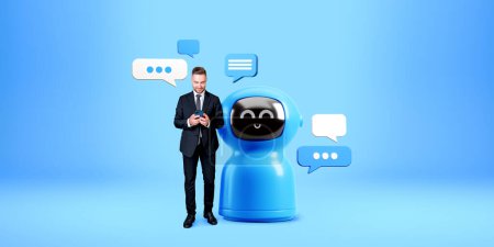 Téléchargez les photos : Bel homme européen en costume debout avec smartphone près de grand robot AI intelligence artificielle bleu chat avec des bulles de parole sur fond bleu. Concept d'apprentissage automatique - en image libre de droit