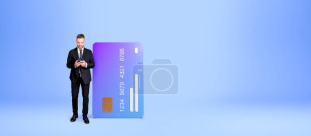 Foto de Joven europeo con teléfono inteligente de pie cerca de la tarjeta de crédito grande sobre fondo azul espacio de copia. Concepto de banca en línea y comercio electrónico - Imagen libre de derechos