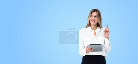Téléchargez les photos : Joyeux jeune femme européenne tenant un ordinateur tablette et debout avec le doigt pointant vers le haut sur fond bleu de l'espace de copie. Concept de bonne idée et de brainstorming - en image libre de droit