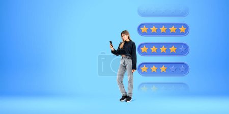 Photo for Young European woman with smartphone and headphones standing over blue copy space background with five stars rating. Concept of product and services evaluation - Royalty Free Image