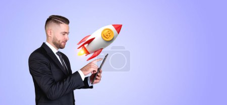 Foto de Joven empresario europeo con smartphone de pie sobre fondo púrpura con cohete bitcoin. Concepto de criptomoneda - Imagen libre de derechos