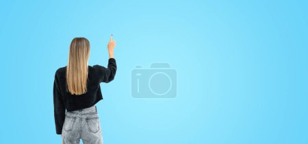 Téléchargez les photos : Vue arrière de la jeune femme en vêtements décontractés en utilisant un écran virtuel sur fond d'espace de copie bleue. Concept de publicité - en image libre de droit