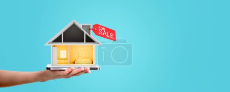 Téléchargez les photos : Main de femme tenant smartphone avec maison à vendre sur fond bleu. Concept d'achat immobilier - en image libre de droit