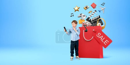Foto de Niño con teléfono inteligente de pie cerca de una gran bolsa de compras con iconos de compras en línea sobre fondo azul. Concepto de comercio electrónico y transacción monetaria - Imagen libre de derechos