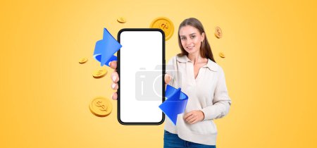 Photo for Smiling woman showing mockup smartphone display, blue arrow with coins falling on yellow background. Concept of refund and digital payment, cashback and money return - Royalty Free Image