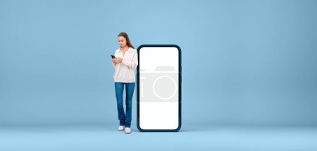 Photo for Young woman standing near large mockup copy space phone screen, blue wide format background. Concept of online network, social media, mobile app and internet communication - Royalty Free Image