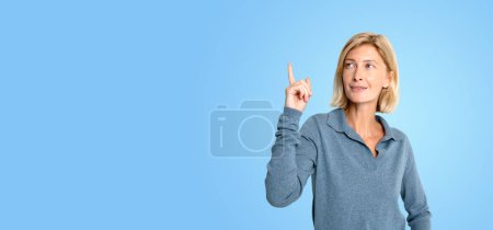 Photo for Portrait of cheerful young European woman pointing upwards with index finger standing over blue background. Concept of planning and bright idea - Royalty Free Image