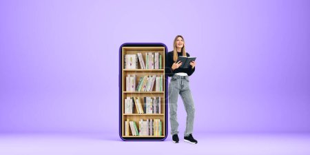 Photo for Dreaming and inspired woman standing with notebook in hands, full length on empty copy space background. Big smartphone screen with books on shelf. Concept of e-learning and digital library - Royalty Free Image