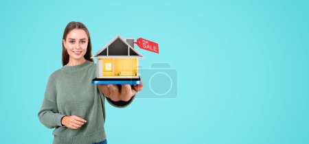 Photo for Smiling woman realtor holding out a small house, red tag for sale. Empty copy space background. Concept of real estate, buy a new apartment, relocation, agent and good offer - Royalty Free Image