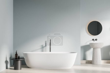 Téléchargez les photos : Intérieur de la salle de bain élégante avec des murs bleu clair, sol en béton, baignoire blanche confortable et confortable évier autoportant avec miroir rond suspendu au-dessus. Rendu 3d - en image libre de droit