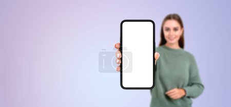 Foto de Retrato borroso de una joven europea sonriente mostrando un teléfono inteligente con pantalla simulada de pie sobre el fondo del espacio de copia púrpura. Concepto de anuncio de aplicaciones y sitios web - Imagen libre de derechos