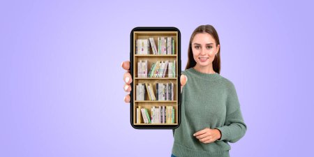 Téléchargez les photos : Portrait d'une jeune étudiante européenne souriante montrant un smartphone avec une bibliothèque numérique debout sur fond d'espace de copie violet. Concept d'apprentissage et de lecture en ligne - en image libre de droit
