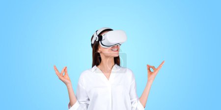 Photo for Cheerful and smiling young woman wearing white shirt and meditating using immersive metaverse interface. Wellbeing and relaxation. Focus on wellness and wearable technology. VR AR mindfullness - Royalty Free Image