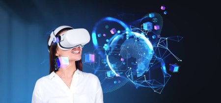 Positive junge Frau mit VR-Headset erkundet den Cyberspace und metaverse. Hologramm des Planeten mit Netzwerksymbolen. Unscharfer blauer Hintergrund. Wearable Geräte der neuen Generation. AR-Technik