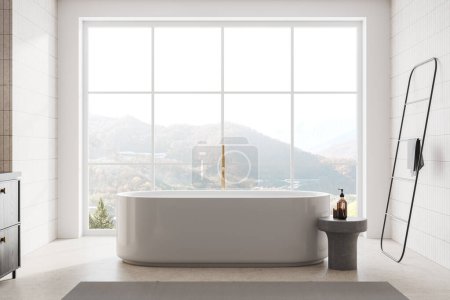 Téléchargez les photos : Salle de bain intérieure de l'hôtel blanche et carrelée avec baignoire, échelle porte-serviettes et table avec accessoires au sol en béton. Fenêtre panoramique sur campagne. rendu 3D - en image libre de droit