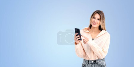 Photo for A smiling woman holding a smartphone, photographic, on a light blue background, conveying a concept of communication - Royalty Free Image