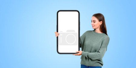 Téléchargez les photos : Une femme tenant une grande maquette vierge de smartphone avec un écran blanc, sur fond bleu, concept de présentation de la technologie - en image libre de droit