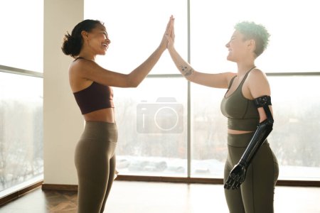 Téléchargez les photos : Jolie fille avec un bras prothétique donnant haute-cinq à l'instructeur pendant qu'ils s'entraînent dans la salle de gym - en image libre de droit