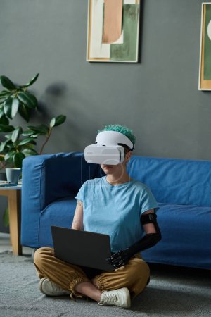Photo for Young woman with prosthetic arm in VR glasses sitting on the floor in the room and using laptop - Royalty Free Image
