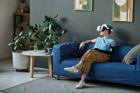 Photo for Young girl with disability watching virtual reality in glasses while sitting on sofa in the living room - Royalty Free Image