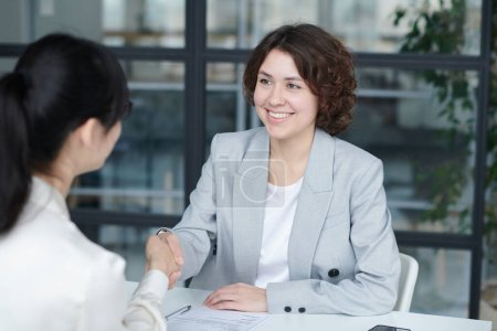 Foto de Sonriente joven gerente estrechando la mano con el empleado para conseguir su trabajo, que se sientan en la mesa durante la reunión de negocios - Imagen libre de derechos