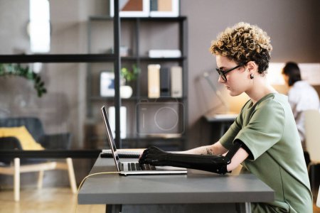 Jeune femme avec un bras prothétique se concentrant sur son travail sur ordinateur portable au bureau pendant la journée de travail