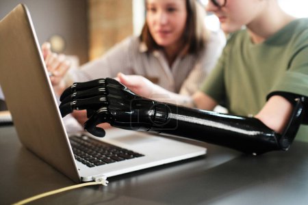 Photo for Close-up of woman with prosthesis arm pointing at monitor of laptop and discussing with her colleague at office desk - Royalty Free Image