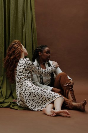 Photo for Young white woman bonding her black girlfriend while they sitting on floor against the brown background - Royalty Free Image