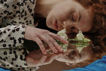 Foto de Mujer joven acostada con los ojos cerrados y tocando la flor acostada en el espejo mojado - Imagen libre de derechos