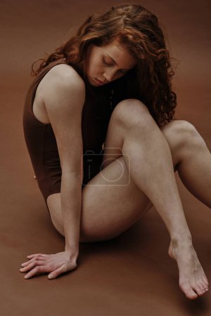 Photo for Young ginger woman with slim body sitting in swimsuit against the brown background - Royalty Free Image