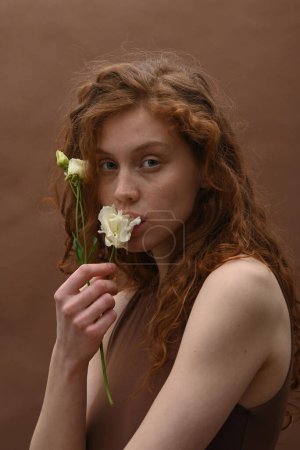 Photo for Portrait of young pretty woman with curly ginger hair holding white flower near her face and looking at camera - Royalty Free Image